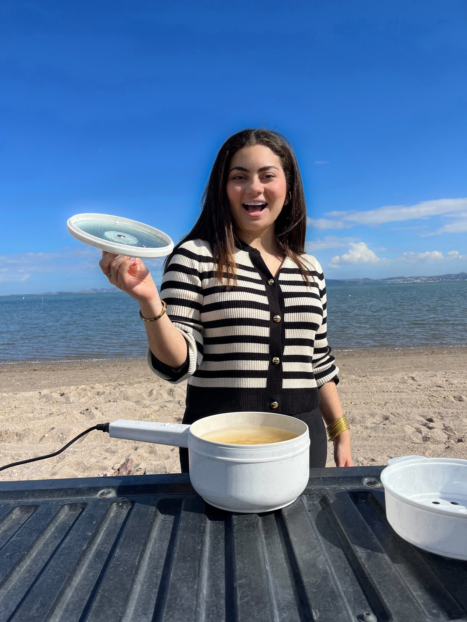 Una mujer alegre sostiene la tapa de una olla eléctrica portátil en la que se está cocinando pasta, sobre una mesa al aire libre con la playa y un cielo azul claro de fondo